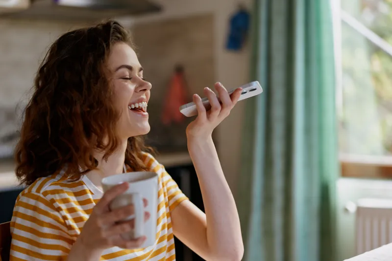 A young woman with brown hair laughs while talking on a smartphone and sipping from a mug inside a bright, cozy home, capturing a candid moment of everyday life.