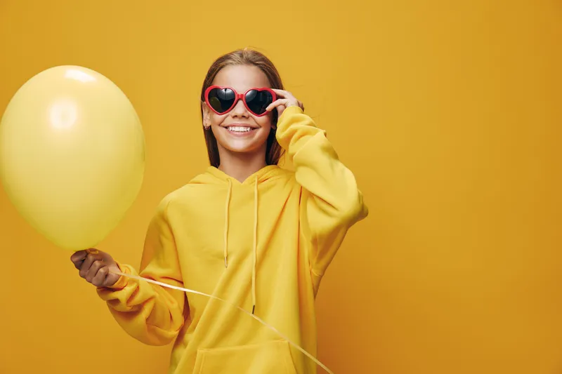Girl in yellow hoodie holding yellow balloon and wearing heart shaped sunglasses standing against yellow background with bright smile and casual pose.