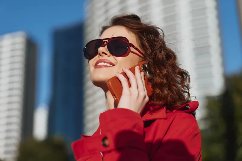 One woman in a red jacket chats on a smartphone outdoors in a sunny city with a smile, tall buildings behind and a clear blue sky, capturing a casual urban moment.