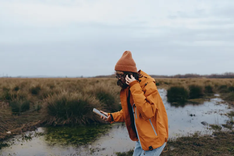 Woman lifestyle, outdoors by river on a cloudy day, wearing orange jacket and knit hat, walking through wet grass, holding a phone, winter mood, travel portrait, alone, soft light