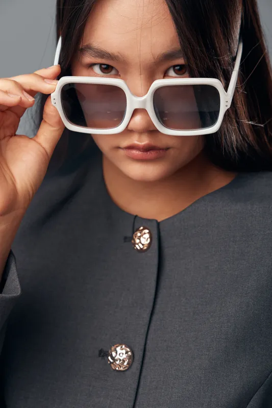 Woman with white sunglasses holding frame and looking at camera in closeup portrait wearing gray jacket with decorative buttons on plain background.
