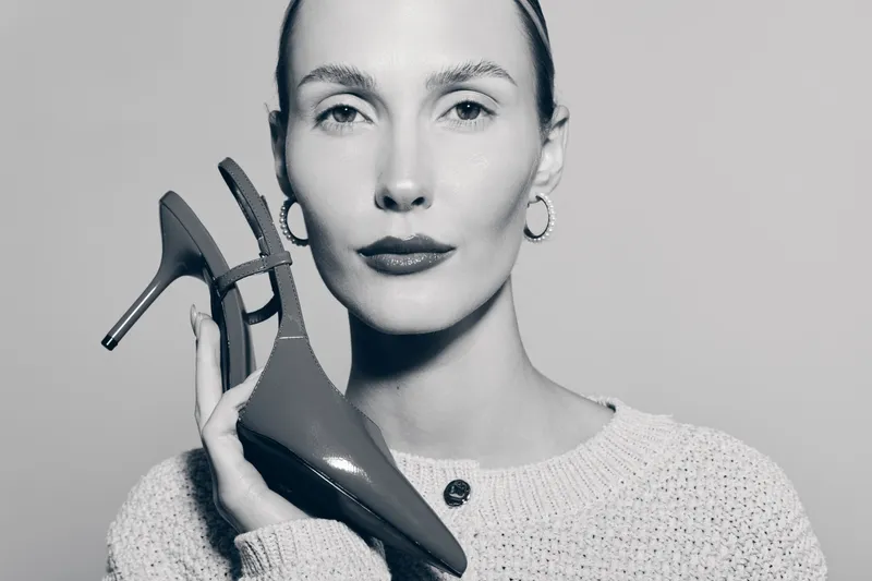 Portrait of a fair skinned white woman holding high heel shoes near her face. Studio shot with soft lighting on neutral background.