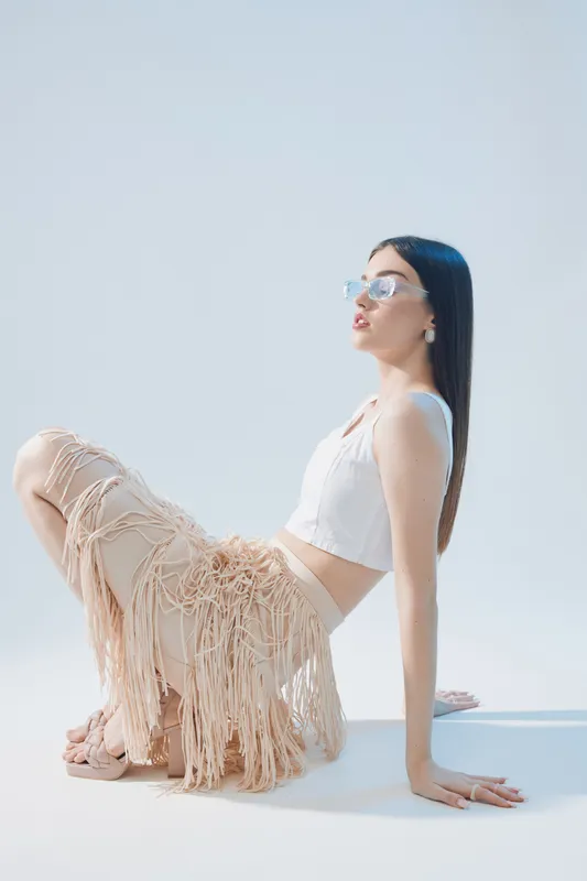 Young woman in stylish fashion outfit with fringe pants and white top poses sitting on floor against white background. Concept of beauty, fashion, and modern style.