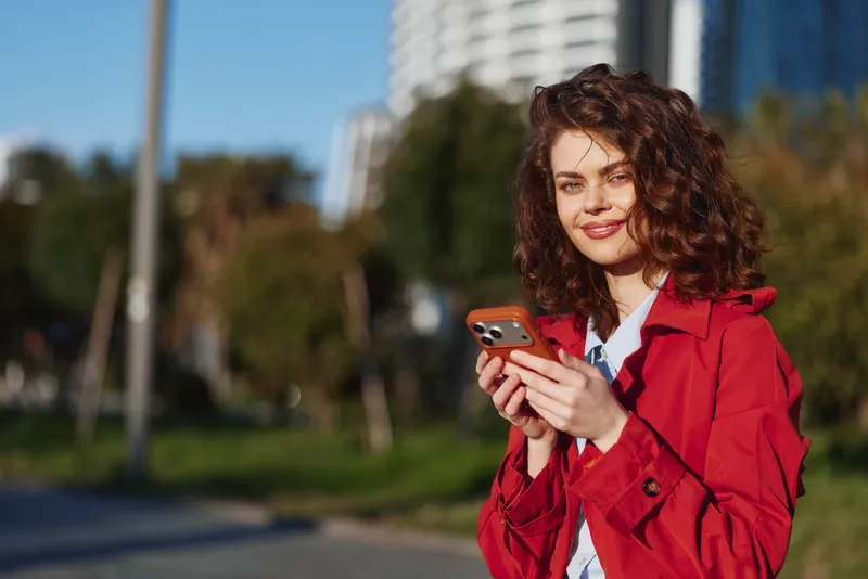 Smiling woman with curly hair wearing a red coat holds a smartphone outdoors in a sunny urban park, clear street background, fashion, lifestyle, and technology concept.