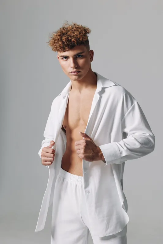 Young man with curly hair wearing white open shirt and pants posing in studio. Fashion model with confident expression and casual style on neutral background.