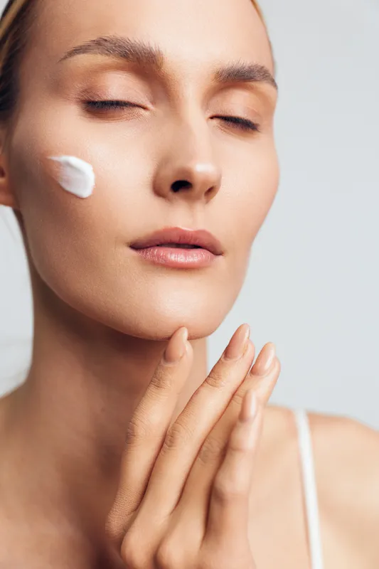 A young woman with closed eyes applies cream on her cheek, focusing on skincare and self-care. The close-up highlights smooth skin and a calm, relaxed expression.