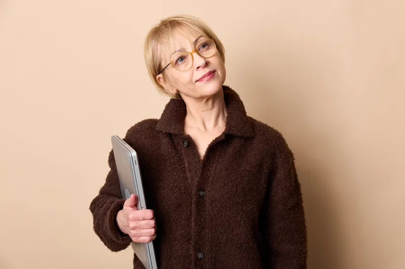 Elderly woman with glasses holding a book, reading pose, studio portrait with beige background, calm expression, brown coat, soft light, attentive posture