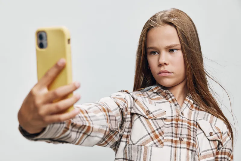 Teen girl with long hair wearing plaid shirt taking selfie by smartphone camera on white background in studio. Portrait, technology and digital communication with serious expression