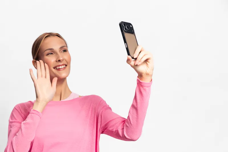 Smiling woman in pink sweater waving hand while taking selfie with smartphone on white background. Concept for communication, technology, social interaction, and happiness.