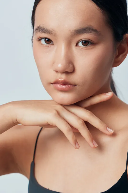 Woman with clear skin posing with hand near face in minimal makeup, natural expression on soft light background for beauty portrait and skincare concept.