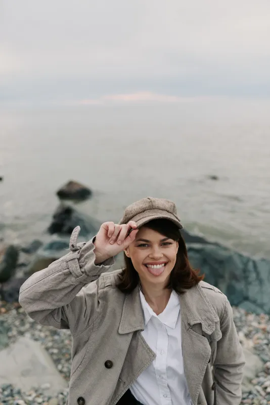 Smiling young woman in a beige coat and cap stands on rocky shore near calm sea. She touches her hat and looks happy. Concept of nature, travel, and outdoor lifestyle.