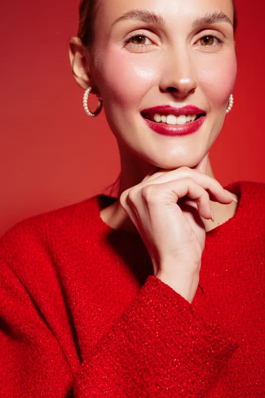 Close-up of smiling woman with red lipstick and earrings wearing red sweater against red background. Concept of beauty, fashion, confidence, and vibrant style in studio lighting.
