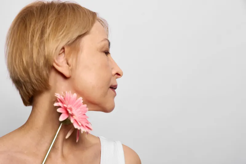 Woman with short blonde hair holds a pink flower to her neck, side profile closeup. Beauty and wellness concept with spa relaxation mood, soft white minimal calm background.