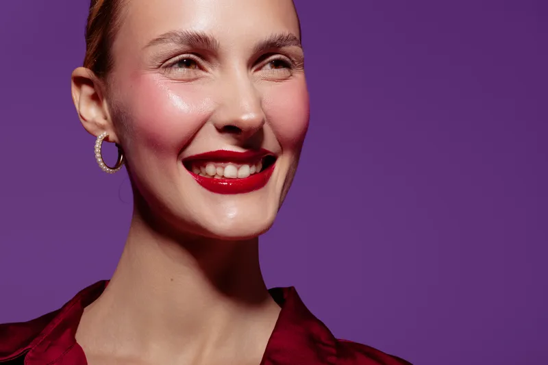 Smiling woman with red lipstick and hoop earrings poses against purple background. Close-up portrait of happy female model with shiny skin and confident expression in studio lighting.