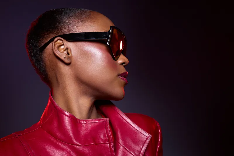 Profile of elegant African American woman with dark skin wearing large sunglasses and red leather jacket. Studio portrait with dramatic lighting on dark purple background. Fashion concept.