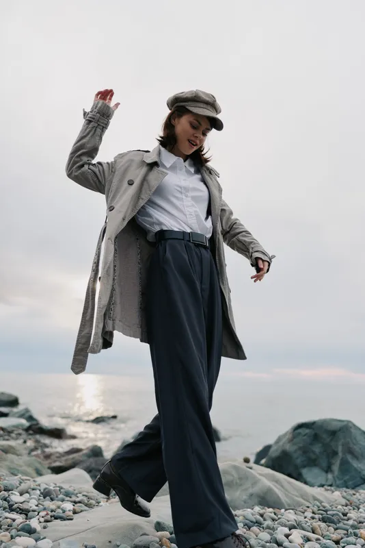 Stylish woman in long coat and cap walks on rocky beach near calm sea under cloudy sky. Casual fashion outfit with wide pants and white shirt creates relaxed and elegant mood outdoors.
