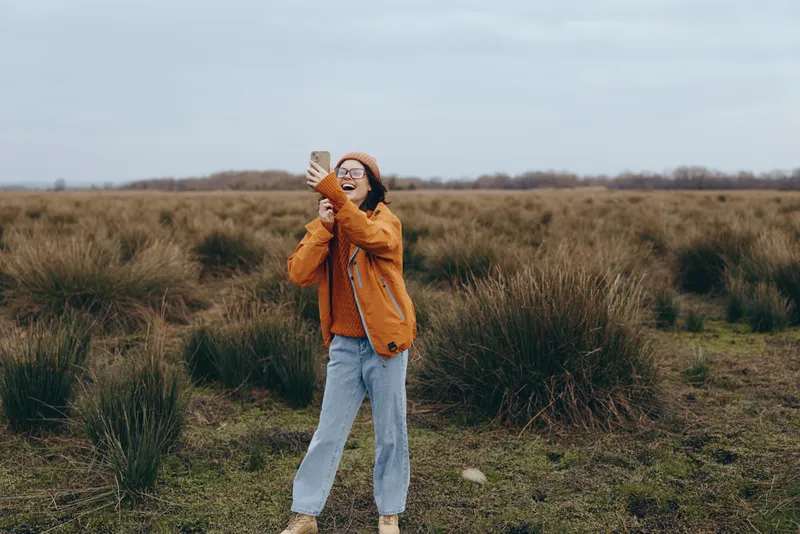 Smiling woman lifestyle outdoors, standing in an autumn field, orange coat, hat in wind, soft overcast light, grass background, casual portrait