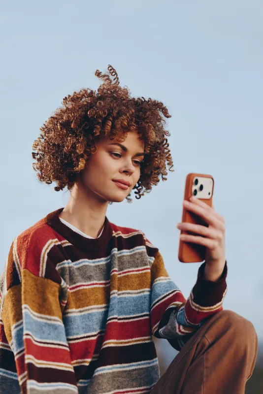Woman lifestyle selfie, curly hair, smartphone portrait outdoors, sky background, casual striped sweater, natural makeup, relaxed smile, holding phone, daylight, travel mood, urban