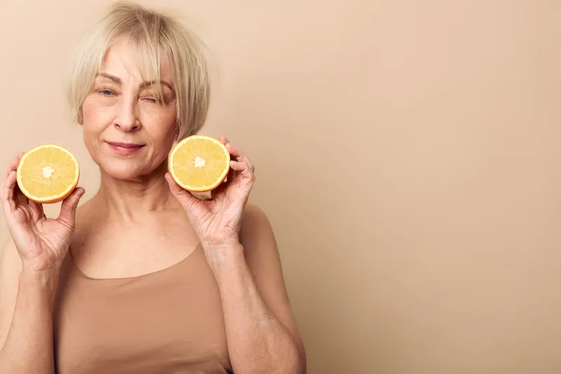 Smiling mature woman with short blonde hair holds two halves of fresh orange near face on beige background. Concept of healthy lifestyle, natural vitamin, wellness, and positive aging.
