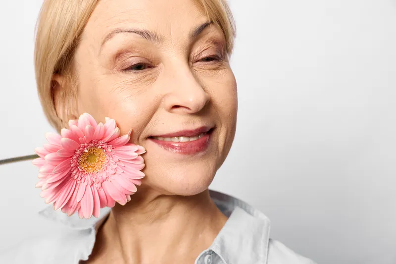 Senior woman portrait smiling softly while holding a pink flower near her cheek, skincare concept, bright studio background, calm happiness and wellbeing.