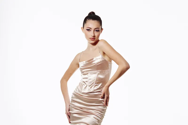 Elegant young woman poses confidently in a shiny champagne satin dress with thin straps against a white background. Fashion concept with a modern beauty and style vibe.