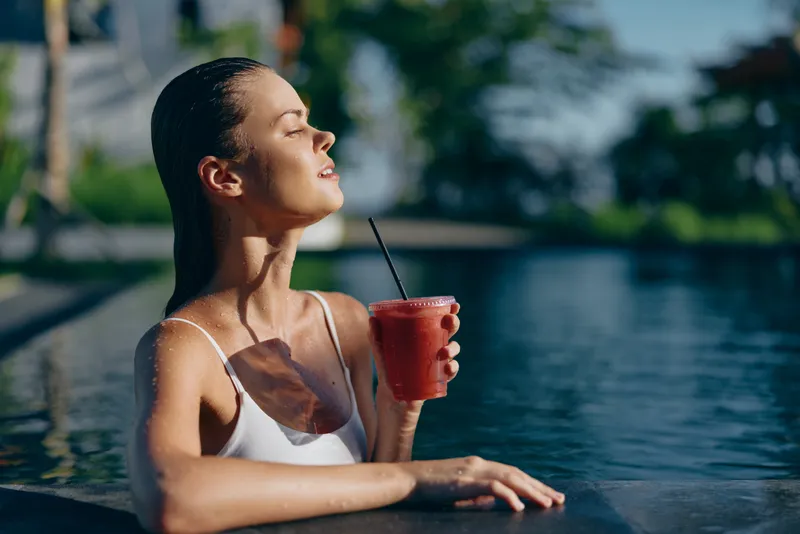 Young woman in white swimsuit relaxes at outdoor pool edge holding red smoothie with straw, eyes closed, enjoying sunny warm day with fresh beverage and calm atmosphere.