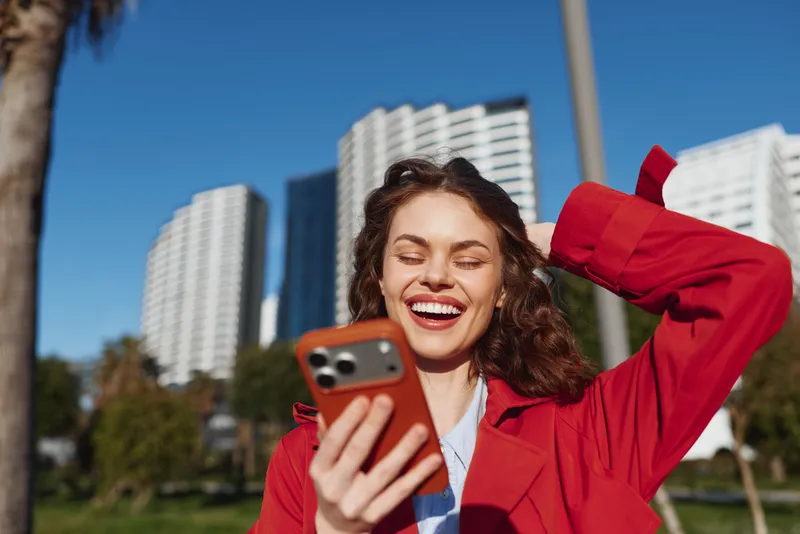 Young brunette woman with curly hair smiles joyfully while taking a selfie outdoors in a red coat against modern city buildings on a sunny day.