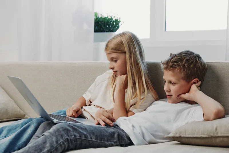 Two children lying on a sofa using a laptop together in a bright living room. The girl is focused on the screen while the boy relaxes, illustrating technology and family time.