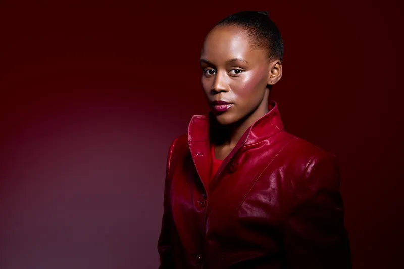 Confident black woman with dark skin wearing a red leather jacket poses against a red gradient background. Studio portrait captures elegance, fashion, and empowerment concepts.