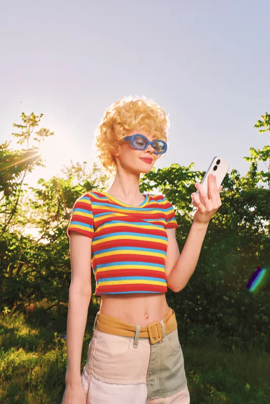 Young woman with curly blonde hair wears a striped croptop and blue sunglasses while taking a selfie outdoors on a sunny day, embracing a carefree retro vibe and warmth through film.