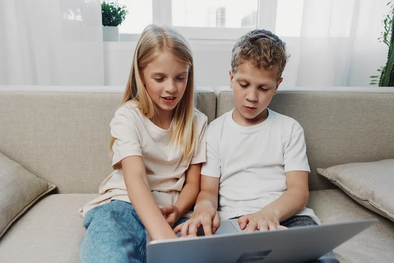 Two children sitting on a sofa using a laptop together in a bright living room. Casual clothing and focused expressions suggest learning or entertainment activity.