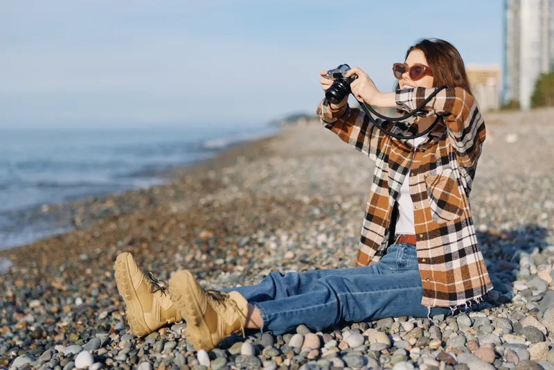 Photographer woman on a rocky beach captures a coastal scene with camera, wearing plaid coat, jeans and boots, enjoying outdoor photography under sunlight by the sea and distant buildings