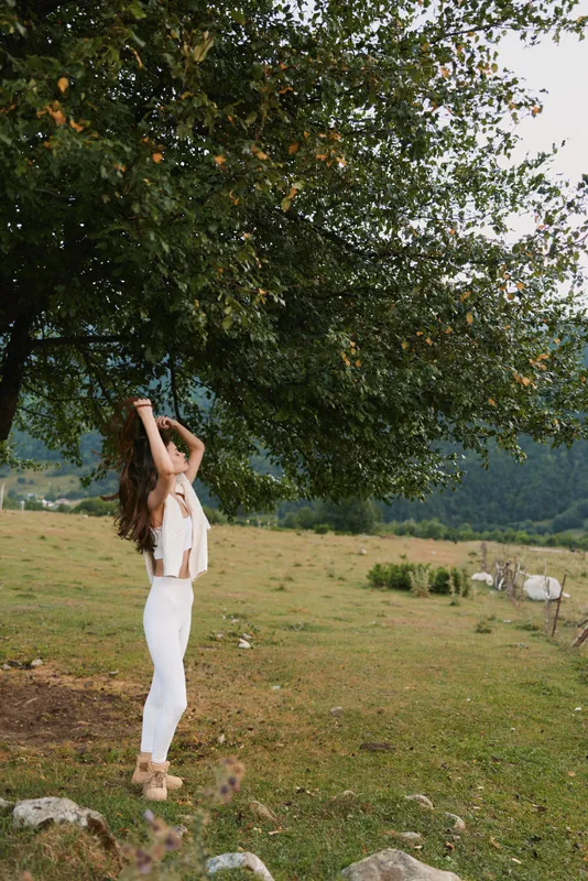 Young woman in white clothes stretching under a large tree in a green field with mountains in the background. Concept of lifestyle, nature, and relaxation outdoors.