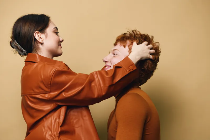 Young couple, woman and man in brown clothes hugging and looking at each other with smiles on beige background. Casual clothing, intimacy, affection and relationship with happy emotions.