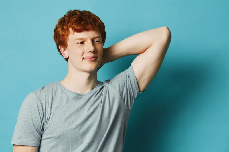 Young man with red hair wearing gray t shirt standing relaxed with hand behind head against blue background. Casual male portrait with confident expression and soft lighting.