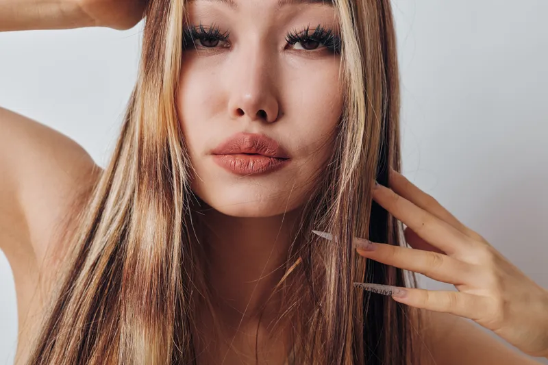 Portrait of young woman with long blonde hair and natural makeup looking at camera, closeup on face with hand touching hair and neutral background.