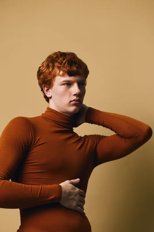 Young redhair man in brown turtleneck sweater posing with hand behind neck looking sideways against brown background in studio portrait with soft lighting and calm expression.