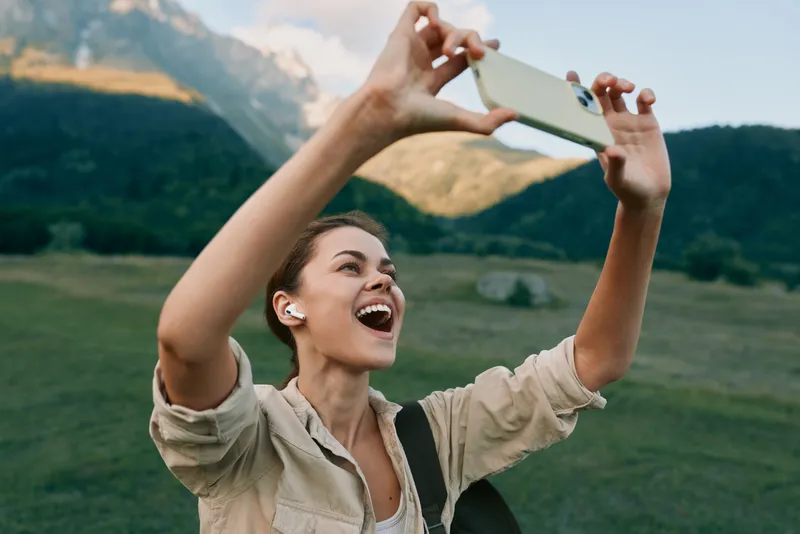 Young woman with earbuds takes a joyful selfie outdoors in a green mountainous landscape during daylight, expressing happiness and freedom with a wide smile.