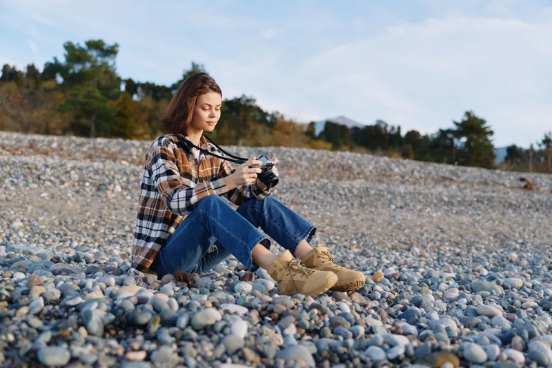 Woman photographer with camera on a pebble beach, casual plaid shirt and boots, focused on framing a shot in outdoor nature by the sea