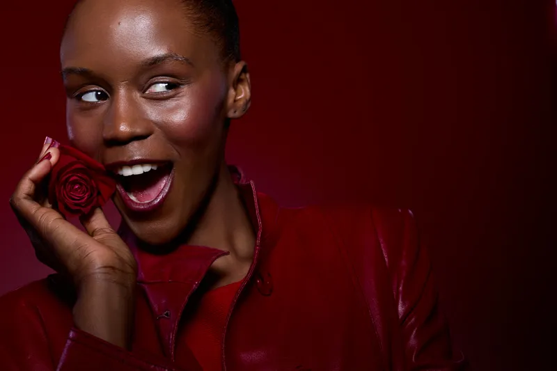 Closeup portrait of a smiling dark skinned African woman holding a red rose near her face. She wears a red jacket and looks to the side on a deep red background. Concept of beauty and joy.