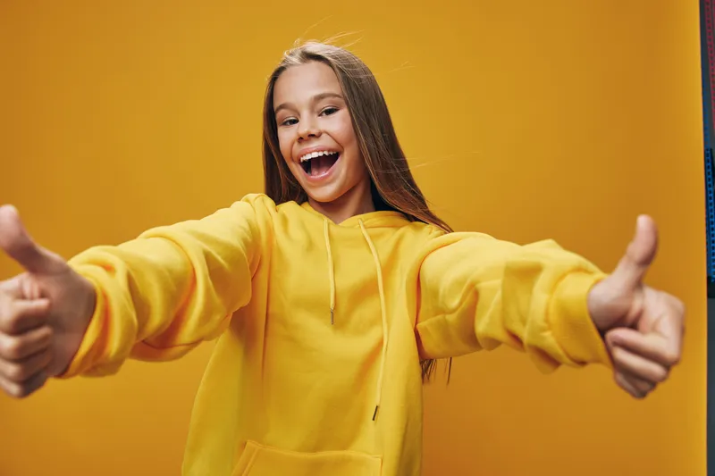 Girl in yellow hoodie showing thumbs up with smile on orange background. Young woman expressing positive emotion and happiness with casual clothing and long hair.