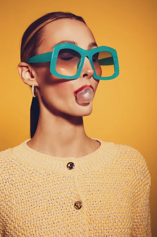 Young woman with large turquoise sunglasses blows a bubblegum bubble. She wears a yellow knitted sweater and a matching headband against a solid yellow background. Fashion and fun concept.