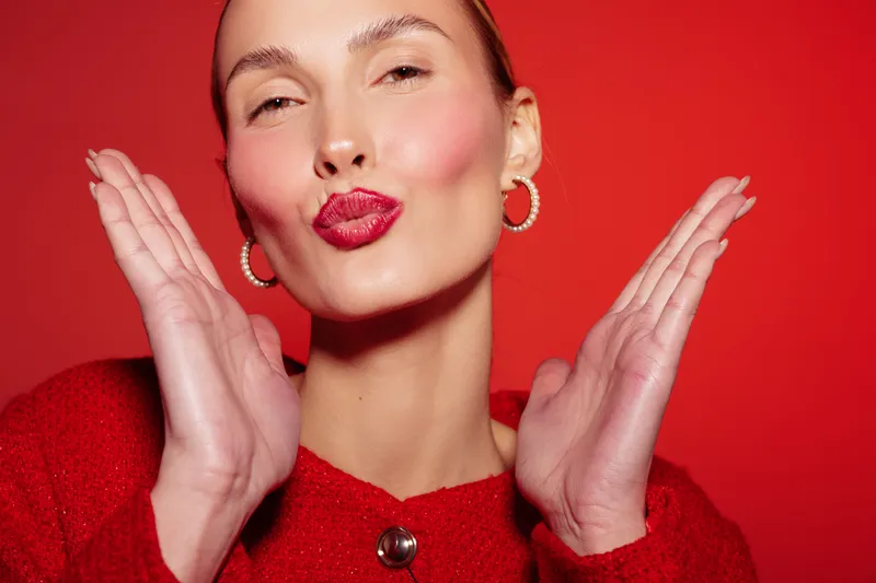 Young woman with red lipstick and hoop earrings blows a kiss with both hands near her face on a red background. Concept of beauty, fashion, and playful expression.