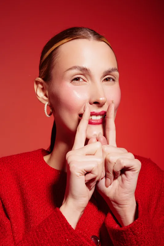 Smiling woman with clear skin and red lips gestures with fingers near mouth on red background. Concept of beauty, skincare, expression, and positive mood in studio lighting.