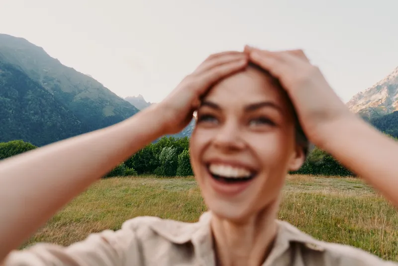 Happy woman with hands on head smiling outdoors in nature with mountains and green field in background. Concept of lifestyle, joy, freedom, and natural beauty.