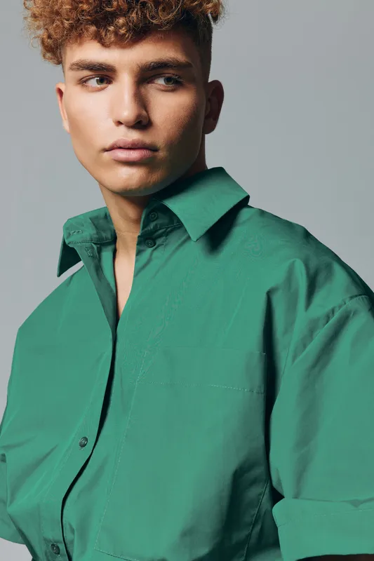 Young man with curly hair wearing green shirt looking sideways with serious expression on gray background. Portrait of male model in casual clothing with natural light and soft shadows.