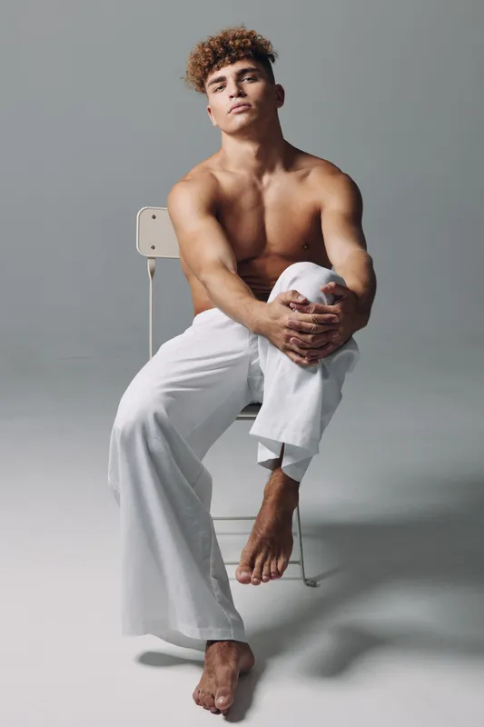 Young muscular man sitting on chair with bare torso and white pants, posing confidently in studio with neutral background and relaxed posture.