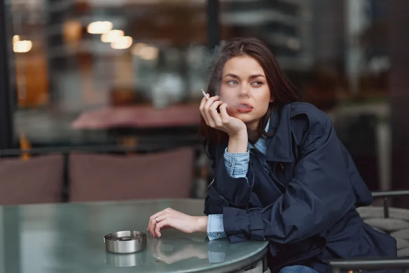 Young woman with dark hair sits at a cafe table, resting her chin on her hand beside an ashtray, lost in thought and distant lights glow softly around her.