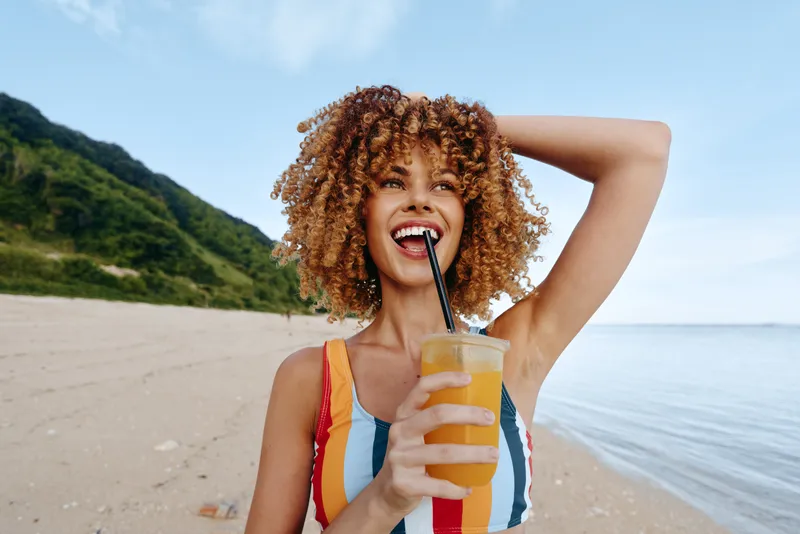 Young woman with curly hair wearing a striped swimsuit stands on a sunny beach, drinking from a cup with a straw.