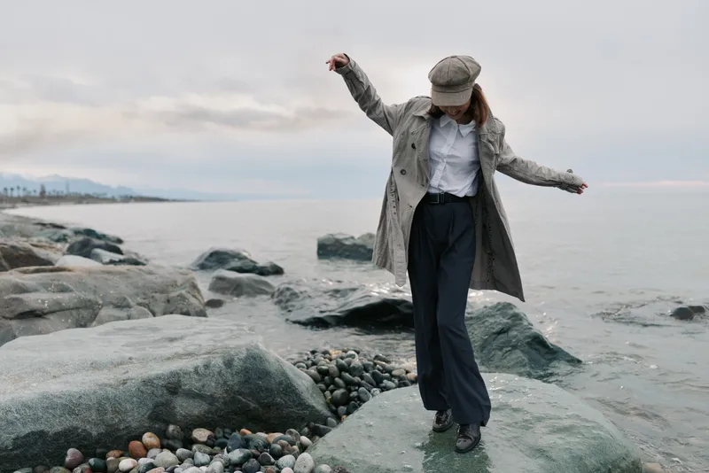 Woman in stylish coat and cap balances on rocky shore near calm sea under cloudy sky. Outdoor fashion, leisure, and nature concept with casual autumn outfit and relaxed mood.